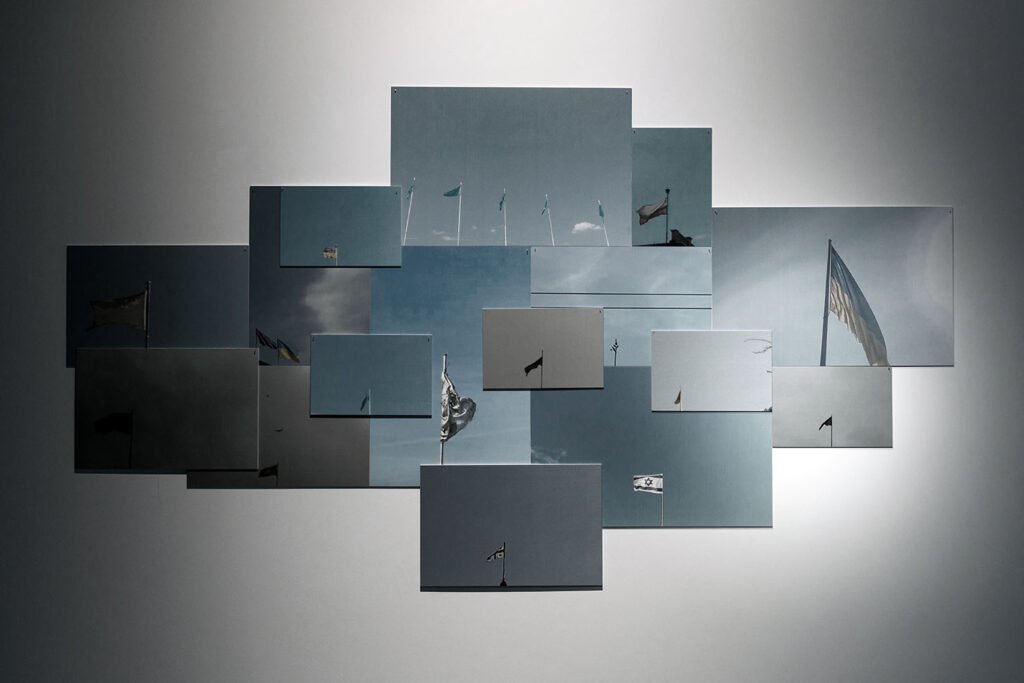 Gęsta horyzontalna kompozycja prostokątnych fotografii, ułożonych bez przerw i częściowo nachodzących na siebie. Każda przedstawia niebo z pojedynczą flagą na maszcie. Różne formaty i odcienie błękitu i szarości tworzą nieregularną, warstwową strukturę.