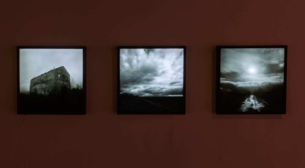 Na ciemnobrązowej ścianie wiszą trzy kwadratowe lightboxy z czarno-białymi fotografiami w czarnych ramach. Pierwsza przedstawia ruinę kamiennego budynku na wzgórzu, druga - dramatyczne niebo nad pustym polem, trzecia - błotnistą drogę w świetle słońca przysłoniętego chmurami. Kompozycja tworzą melancholijny, filmowy nastrój.