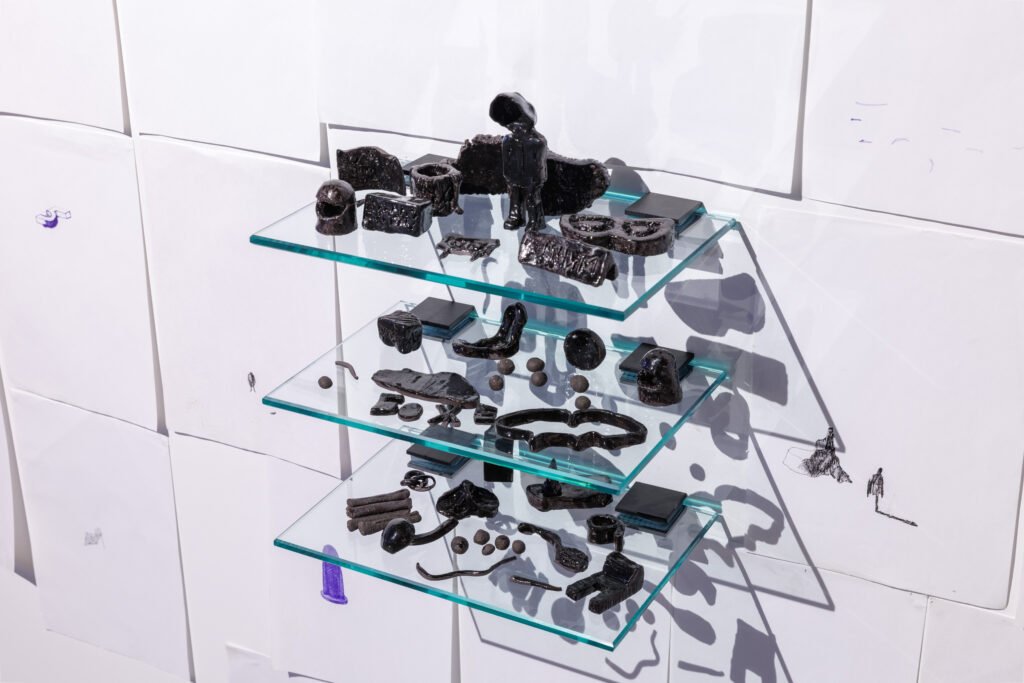 Na białej ścianie, pokrytej kartkami papieru, zawieszono trzy przezroczyste szklane półki. Na każdej z nich stoją liczne małe, czarne obiekty rzeźbiarskie o  nieregularnych kształtach. Ostre światło rzuca wyraziste cienie form na ścianę.